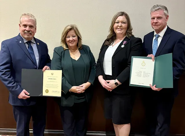 Four individuals are standing side by side, dressed in formal business attire, in a room with a neutral-toned wall and patterned carpet. From left to right: John Jordan, MPP for Lanark-Frontenac-Kingston, holding a certificate; Lisa Thompson, Minister of Rural Affairs; Christa Lowry, Mississippi Mills Mayor and ROMA Chair, wearing a King Charles III Coronation Medal on her lapel; and Steve Pinsonneault, Parliamentary Assistant to the Minister of Rural Affairs, also holding a certificate. The group is gathered to celebrate the recognition of Mayor Lowry with the King Charles III Coronation Medal during the ROMA Conference.
