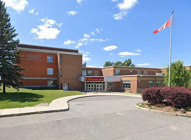 A large, red-brick high school building with a circular driveway in front. A Canadian flag flies from a pole on the right, and the sign above the entrance reads 