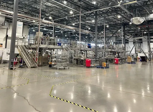 Large, brightly lit indoor cannabis sales production facility with multiple levels of stainless steel equipment, walkways, and yellow safety lines on the concrete floor.