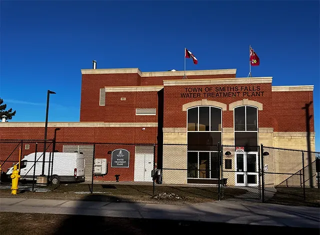 Exterior photo of the Town of Smiths Falls Water Treatment Plant, a brick building with flags flying, on a clear day. A white work van is parked in front of the building, which is surrounded by a chain-link fence. The building's sign mentions a water treatment plant, which is connected to the town's water meter replacement project.